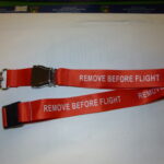 Porta Badge Remove Before Flight di colore rosso da collo stampato, con chiusura cintura sicurezza aereo.